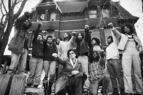 Black and white photo of male and female Black MOVE members standing and raising their fists in front of their house. 